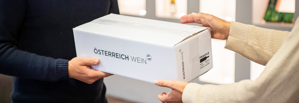 Das Bild zeigt eine Person, welche einer anderen Person einen Karton mit der Aufschrift "Österreich Wein" reicht.